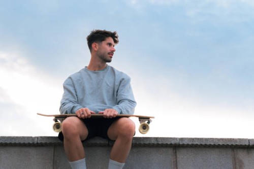 young man sitting with a skateboard in a cloudy day at sunset - fashion photos et images de collection