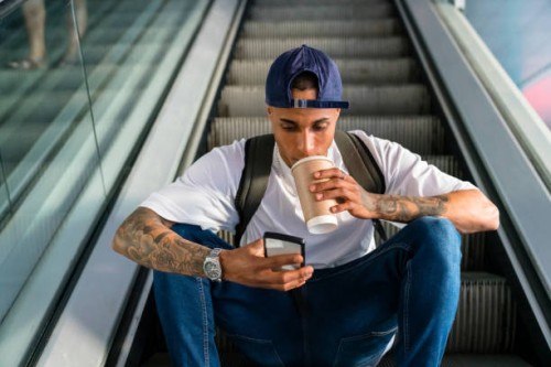 young man with backpack sitting on escalator looking at cell phone while drinking coffee to go - junk food stock pictures, royalty-free photos & images