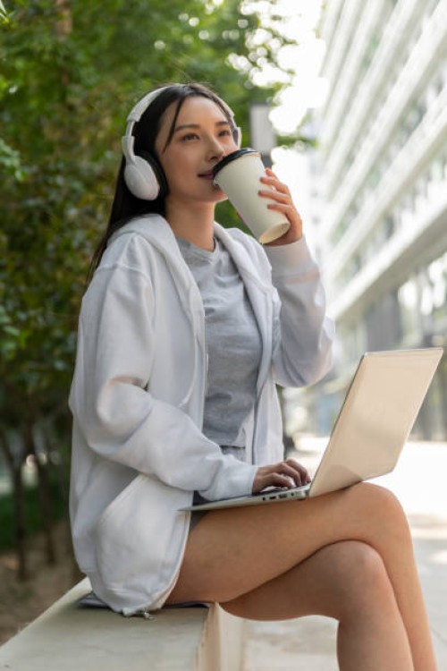 young smiling woman in headphones working on laptop and drinking takeaway coffee while sitting outdoors - junk food stock pictures, royalty-free photos & images