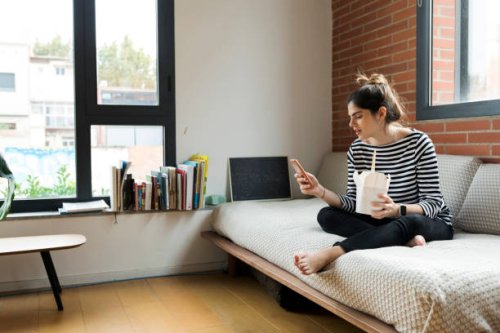 young woman sitting on couch at home having asian takeaway food and using cell phone - junk food stock pictures, royalty-free photos & images