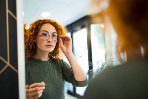 young woman trying on glasses in optical store looking at mirror - fashion stock pictures, royalty-free photos & images