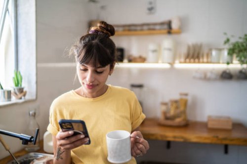 young woman using the mobile phone while drinking coffee or tea at home - coffee stock pictures, royalty-free photos & images