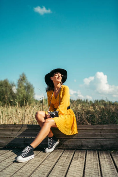 young woman wearing a black hat and yellow dress with an analog camera sitting on wooden boardwalk - fashion photos et images de collection