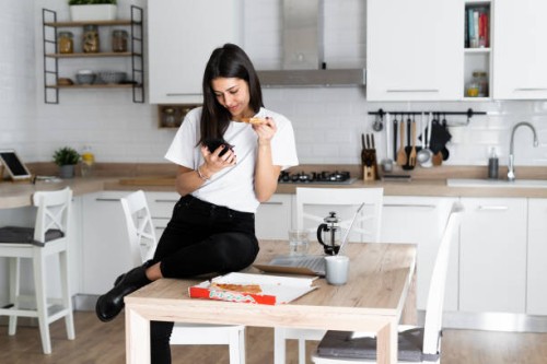 young woman with cell phone eating pizza in kitchen at home - junk food stock pictures, royalty-free photos & images