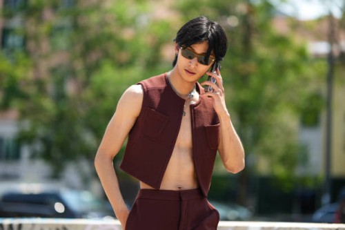Yugo Takano wears brown sunglasses, silver large chain long necklaces, a brown burgundy sleeveless / cropped gilet, matching brown burgundy flared...